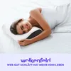 A woman is lying on a bed with a pillow, smiling at the camera. Produktbild-Vorschau