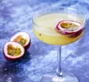 A close-up shot of a passion fruit cocktail in a coupe glass, garnished with a slice of passion fruit. Two passion fruit halves are visible in the background. Podgląd zdjęcia produktu