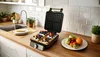 A waffle maker is open and filled with waffles topped with fruit and cream, sitting on a kitchen counter next to a sink and a bowl of fruit. Podgląd zdjęcia produktu