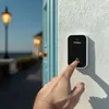 A hand presses a doorbell on a white wall next to a blue door. A street lamp and a building are visible in the background. Náhľad obrázku produktu
