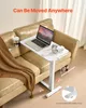 A white mobile laptop table with a laptop, headphones, and mouse on it, placed next to a tan leather sofa with a brown blanket and a cup holder with a cup. Produktbild-Vorschau