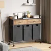 A laundry cart with three fabric bins and two drawers, placed in a room setting with a floor lamp and a shelf. Produktbild-Vorschau