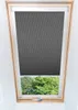 A dark gray roller blind is partially lowered over a skylight, revealing green trees and foliage outside. Voir les images du produit