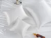 Three white silk pillowcases of different sizes are arranged on a white, creased surface, possibly a bed. A small cup and saucer and a bottle of amber liquid are also visible. Podgląd zdjęcia produktu