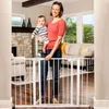 A woman holds a baby while standing in front of a white baby gate in a living room. Voir les images du produit