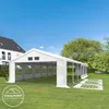 A white party tent is set up on a green lawn in front of a house and trees, under a clear blue sky. Several white ground frame pieces are laid out in front of the tent. Produktbild-Vorschau