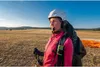 A woman in paragliding gear stands in a field with a paraglider in the background. Náhled obrázku produktu