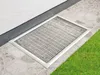 A metal grate cover for a basement window well, placed on a stone patio next to a patch of grass. Produktbild-Vorschau