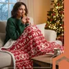 A woman is sitting on a couch, wrapped in a festive red and white blanket, eating a cookie. A Christmas tree is visible in the background. Náhled obrázku produktu