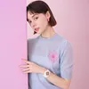 A young woman leans against a pink surface, wearing a light blue sweater with a pink flower design and a white watch. Anteprima dell’immagine prodotto
