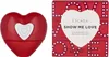 A red heart-shaped perfume bottle and its matching red box are displayed side-by-side against a white background. Produktbild-Vorschau