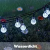 String lights hanging in a garden during rainfall, with flowers in the background. Produktbild-Vorschau