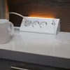 A white corner power strip with three outlets and USB ports is shown on a white countertop, with a mug to the left and a wooden backsplash behind it. An orange light emanates from the top of the power strip. Produktbild-Vorschau