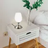 A bedside table with a lamp and a vase of greenery. Produktbild-Vorschau