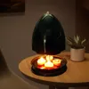 A dark green candle heater with four lit tea lights underneath, placed on a wooden table next to a potted plant. Produktbild-Vorschau