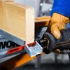 A person wearing a blue shirt and brown gloves is using a black and orange reciprocating saw to cut a wooden beam that is resting on a metal support. Voir les images du produit