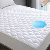 A hand is pouring blue liquid onto a white quilted mattress protector, demonstrating its waterproof feature. Produktbild-Vorschau