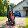 A black and red pressure washer is positioned on a gravel driveway in front of a house and a blue car. Náhled obrázku produktu