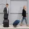 A man and a woman walking with luggage on a sidewalk in front of a textured wall. Podgląd zdjęcia produktu