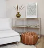 A living room scene with a white sofa, a brown leather pouf, and a shelf with decorative items. Produktbild-Vorschau