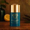 A Versace Eros perfumed deodorant stick is displayed on a wooden surface with a blurred background of a green vase and a lemon slice. Podgląd zdjęcia produktu