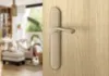 A close-up of a bronze door handle on a wooden door, with a blurred living room in the background. Produktbild-Vorschau