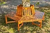 A wooden bench is placed around a tree trunk in a grassy area, with dappled sunlight filtering through leaves. Náhled obrázku produktu