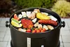 A round charcoal grill with various foods cooking on the grate, including steak, sausage, corn, and vegetables, set up outdoors on a paved patio. Náhled obrázku produktu