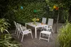 An outdoor dining set with chairs and a table is arranged on a gravel patio, surrounded by lush green foliage and trees. Produktbild-Vorschau