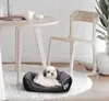 A small dog is resting in a pet bed placed on a fluffy rug next to a wooden chair and a round table. Anteprima dell’immagine prodotto
