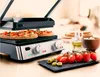 A panini press with two open-faced sandwiches on it, surrounded by fresh ingredients like tomatoes and olives. Podgląd zdjęcia produktu