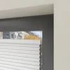 A close-up view of a white pleated blind in a window frame, with a blurred view of buildings and trees outside. Produktbild-Vorschau