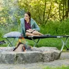 A woman is reading a book while sitting on a camping cot next to a campfire. Náhled obrázku produktu