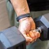 A person's hand is holding a dumbbell. The person is wearing a black fitness tracker on their wrist. Produktbild-Vorschau