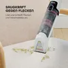 A handheld stain remover is being used to clean a green stain on a light-colored fabric chair. Podgląd zdjęcia produktu
