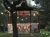 A gazebo decorated with lanterns and string lights in a garden setting at dusk. Podgląd zdjęcia produktu