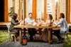 A group of people are having dinner outdoors at a wooden table, with a house in the background. Produktbild-Vorschau