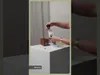 A person is holding a product over a white pedestal with a wooden object and a small device on it. Produktbild-Vorschau