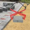 A red paving stone clamp is shown in use, lifting a gray paving stone, with a pile of paving stones in the background. Podgląd zdjęcia produktu