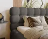 A close-up view of a bed with a dark gray bouclé headboard and pillows, set against a wall with a nature-themed mural. Produktbild-Vorschau