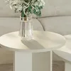 A close-up shot of a round, sand-colored side table with a glass vase of flowers on top, placed in front of a light-colored sofa. Voir les images du produit