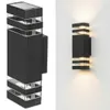 A product shot of a black outdoor wall light with clear glass accents, shown against a white background. To the right, the same light is shown mounted on a white wall, illuminating upwards and downwards. Produktbild-Vorschau