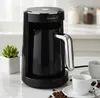 A black coffee maker with a silver handle sits on a marble countertop next to two coffee cups and coffee beans. Produktbild-Vorschau