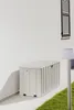 A light gray storage box is placed against a white wall on a paved area with grass to the right. Produktbild-Vorschau