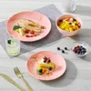 A table setting with pink marble-patterned plates, a glass of water, and fruit salad. Produktbild-Vorschau