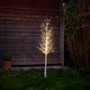 A decorative LED light tree with a birch-like appearance is set up outdoors at dusk, in front of a wooden wall and next to some greenery. Produktbild-Vorschau