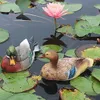 Two decorative duck statues floating on lily pads in a pond with a pink water lily. Produktbild-Vorschau
