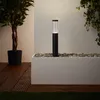 A modern outdoor lamp stands in a planter filled with pebbles, next to a large potted plant and a wooden deck. Produktbild-Vorschau