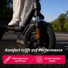 Close-up of an electric scooter's front wheel and deck, with a person's legs visible, outdoors with trees and sky in the background. Produktbild-Vorschau
