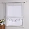 A sheer white Roman blind with a decorative embroidered border hangs on a window. Produktbild-Vorschau